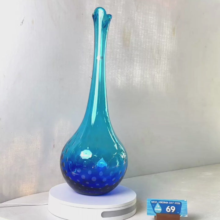 A translucent blue glass vase with a flared neck and dotted pattern, displayed on a white circular base against a light gray wall.
