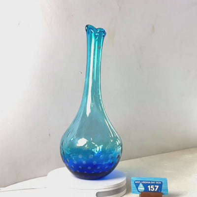A translucent blue glass vase with a flared rim and dotted pattern near the base, displayed on a white circular stand against a plain light gray wall.