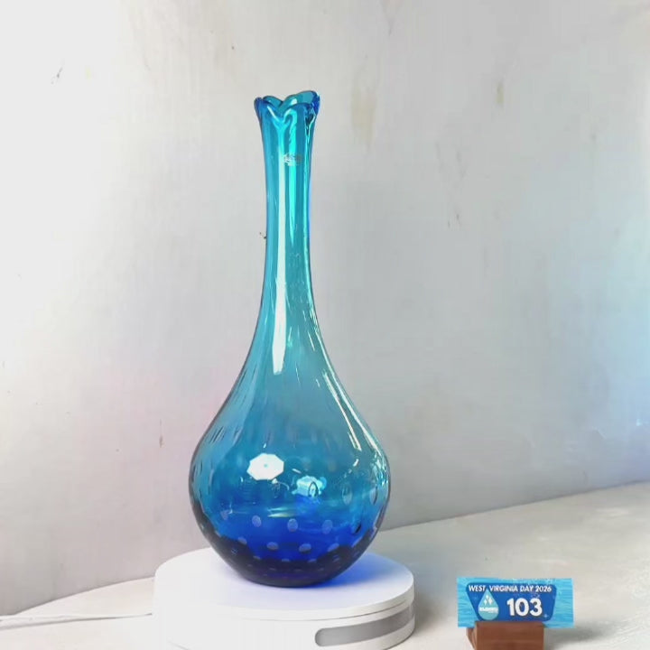 A translucent blue glass vase with a flared rim and dotted pattern near the base, displayed on a white circular stand against a plain light gray wall.