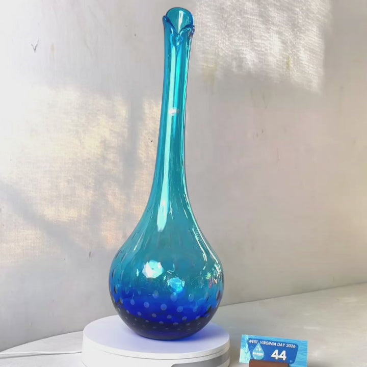 A translucent blue glass vase with a flared neck and dotted pattern, displayed on a white circular base against a light gray wall.