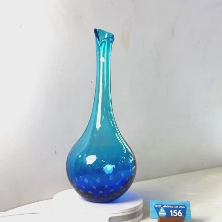 A translucent blue glass vase with a flared rim and dotted pattern near the base, displayed on a white circular stand against a plain light gray wall.