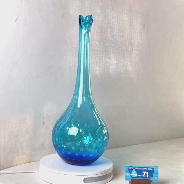 A translucent blue glass vase with a flared neck and dotted pattern, displayed on a white circular base against a light gray wall.