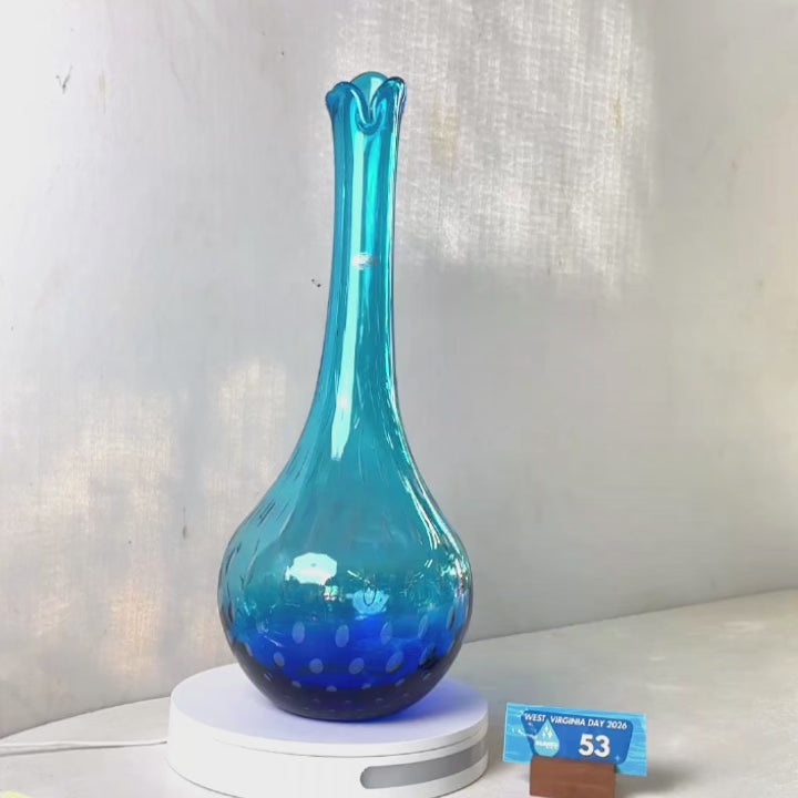A translucent blue glass vase with a flared neck and dotted pattern, displayed on a white circular base against a light gray wall.
