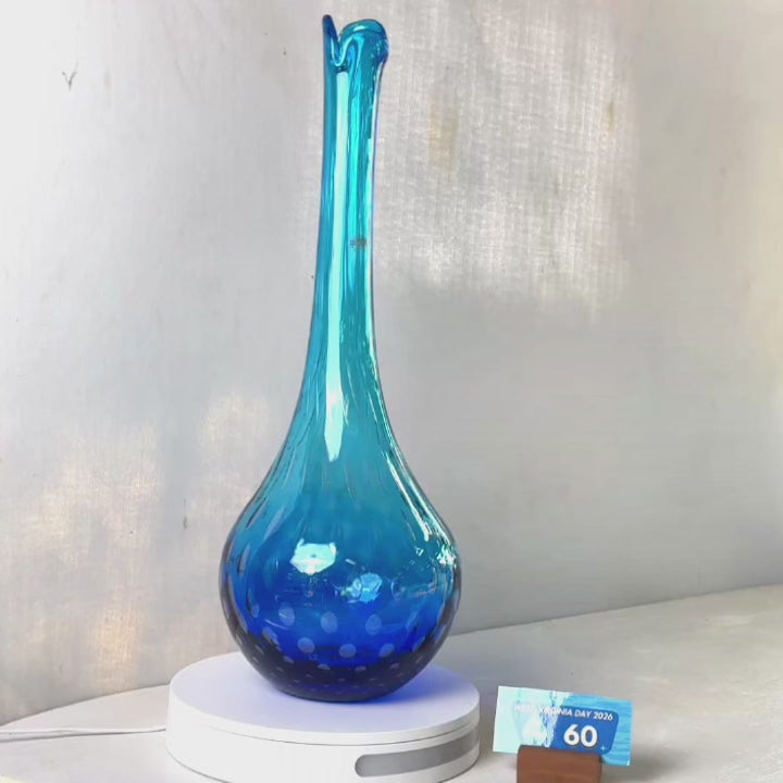 A translucent blue glass vase with a flared neck and dotted pattern, displayed on a white circular base against a light gray wall.