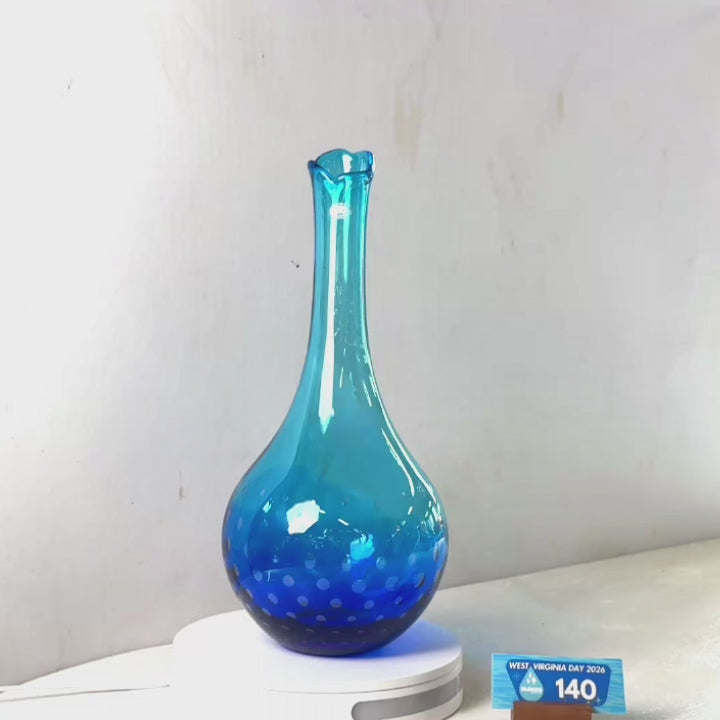 A translucent blue glass vase with a flared rim and dotted pattern near the base, displayed on a white circular stand against a plain light gray wall.