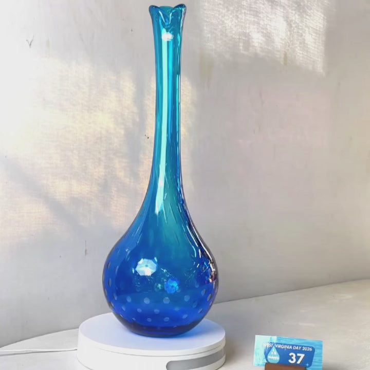 A translucent blue glass vase with a flared neck and dotted pattern, displayed on a white circular base against a light gray wall.