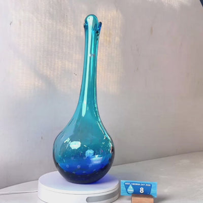 A translucent blue glass vase with a flared neck and dotted pattern, displayed on a white circular base against a light gray wall.