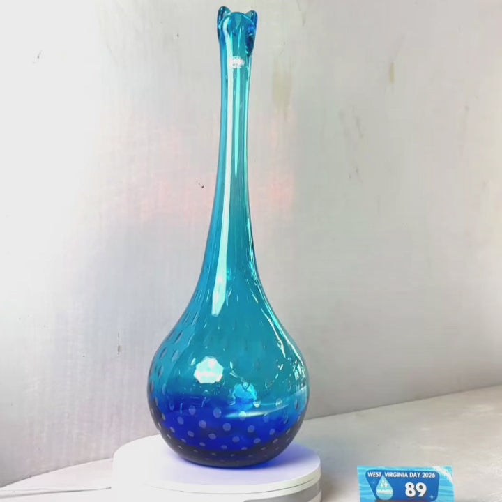 A translucent blue glass vase with a flared rim and subtle white speckled pattern near the base, displayed on a white round scale with a small price tag in front, against a plain light gray wall.