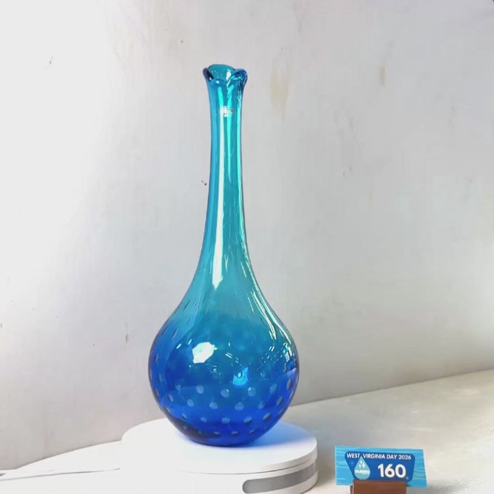 A translucent blue glass vase with a flared rim and dotted pattern near the base, displayed on a white circular stand against a plain light gray wall.