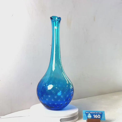 A translucent blue glass vase with a flared rim and dotted pattern near the base, displayed on a white circular stand against a plain light gray wall.