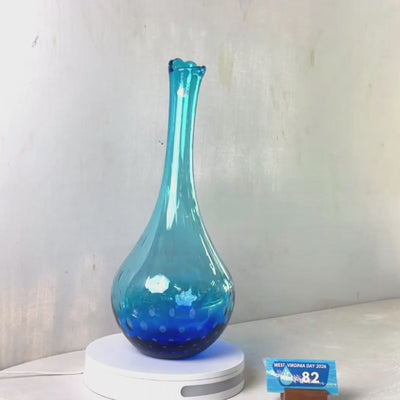 A translucent blue glass vase with a flared rim and subtle white speckled pattern near the base, displayed on a white round scale with a small price tag in front, against a plain light gray wall.