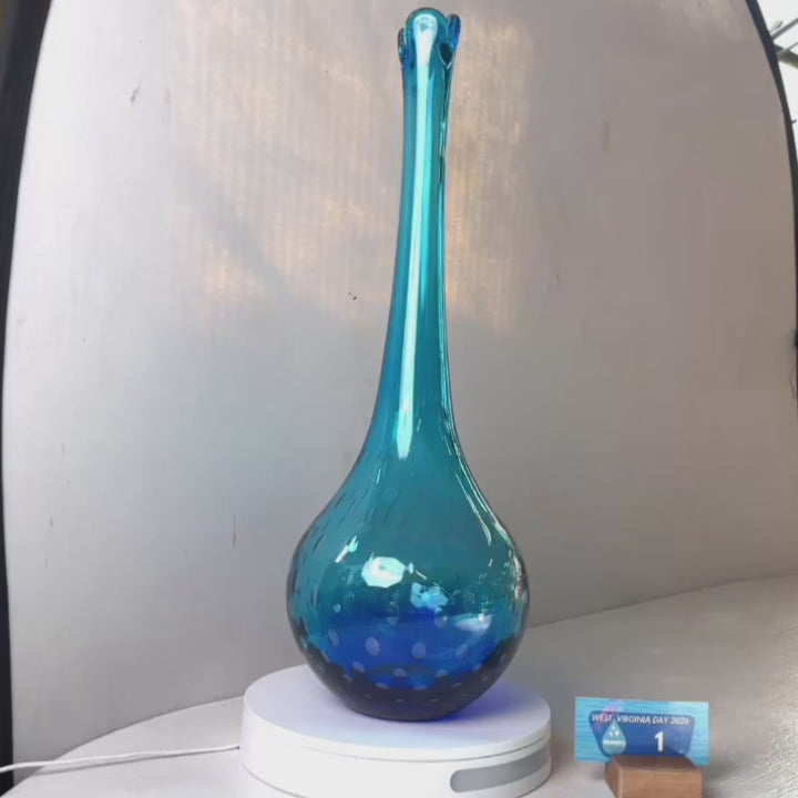 A translucent blue glass vase with a flared neck and dotted pattern, displayed on a white circular base against a light gray wall.