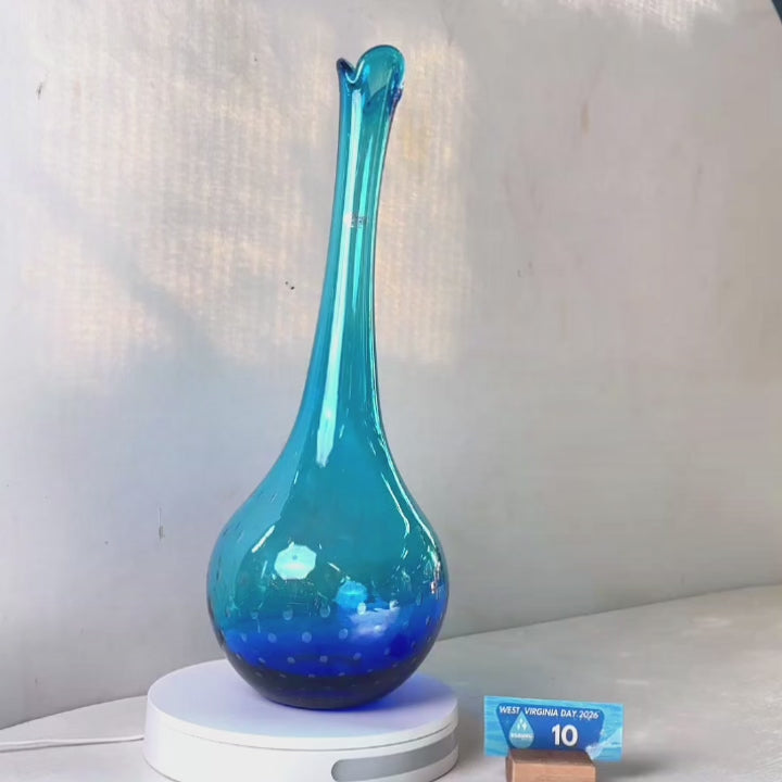 A translucent blue glass vase with a flared neck and dotted pattern, displayed on a white circular base against a light gray wall.