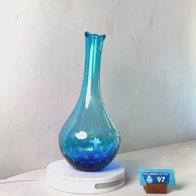 A translucent blue glass vase with a flared rim and subtle white speckled pattern near the base, displayed on a white round scale with a small price tag in front, against a plain light gray wall.