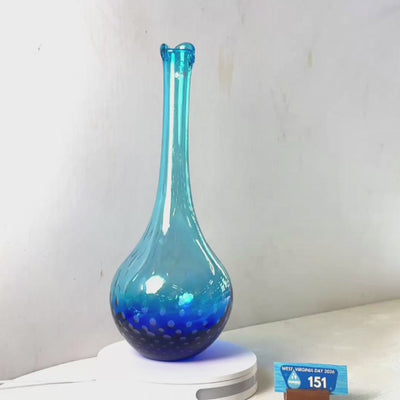 A translucent blue glass vase with a flared rim and dotted pattern near the base, displayed on a white circular stand against a plain light gray wall.