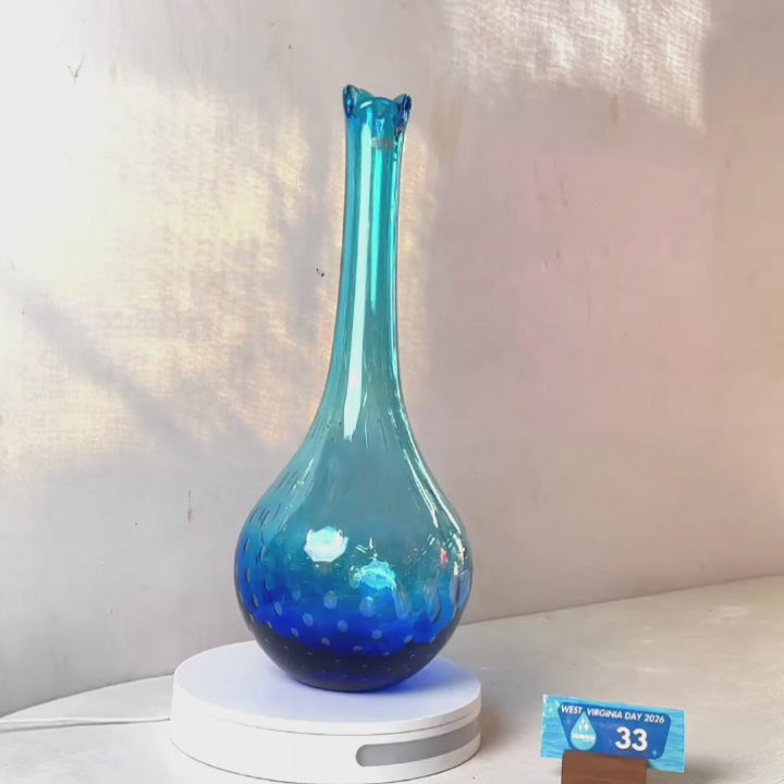 A translucent blue glass vase with a flared neck and dotted pattern, displayed on a white circular base against a light gray wall.