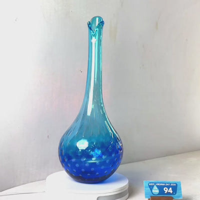 A translucent blue glass vase with a flared rim and subtle white speckled pattern near the base, displayed on a white round scale with a small price tag in front, against a plain light gray wall.