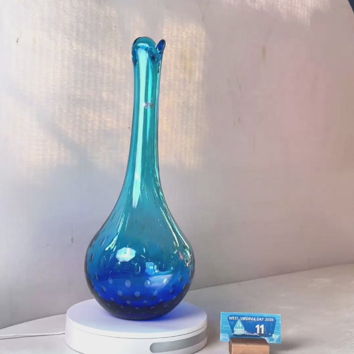 A translucent blue glass vase with a flared neck and dotted pattern, displayed on a white circular base against a light gray wall.