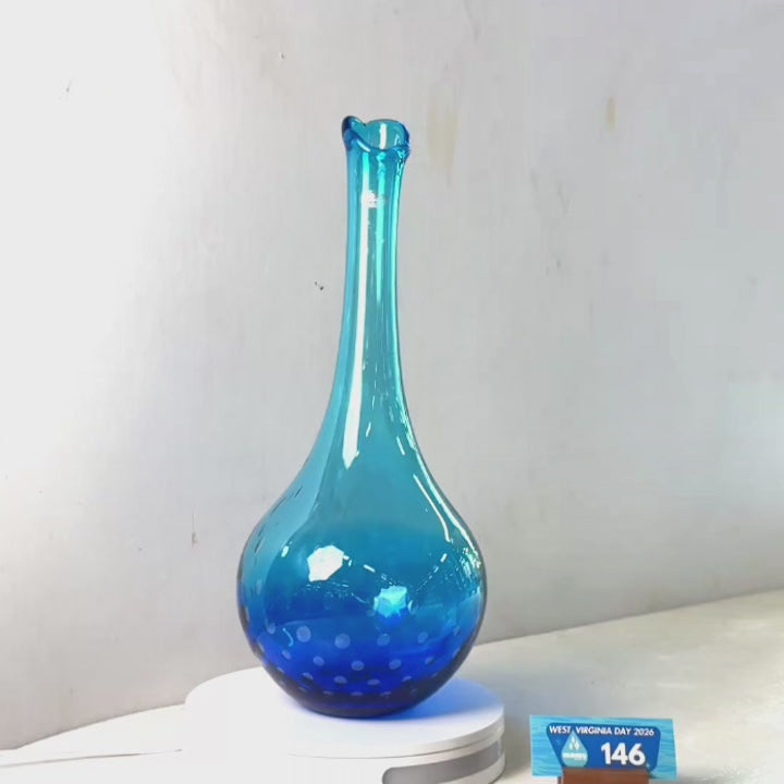 A translucent blue glass vase with a flared rim and dotted pattern near the base, displayed on a white circular stand against a plain light gray wall.