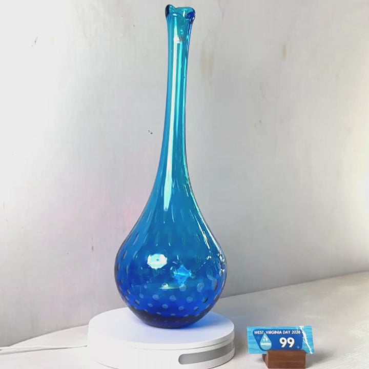 A tall, slender cobalt blue glass vase with a flared neck and small white polka dots, displayed on a white circular light box against a plain light gray wall.