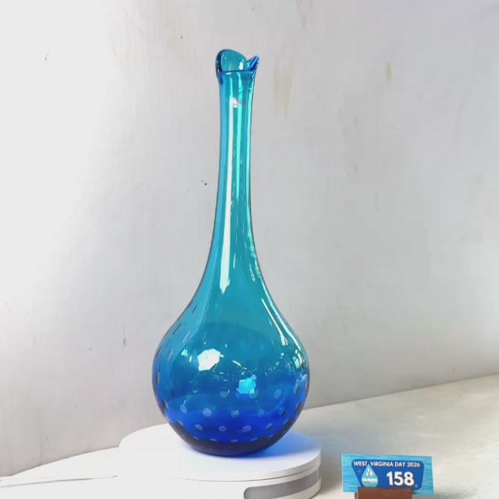 A translucent blue glass vase with a flared rim and dotted pattern near the base, displayed on a white circular stand against a plain light gray wall.