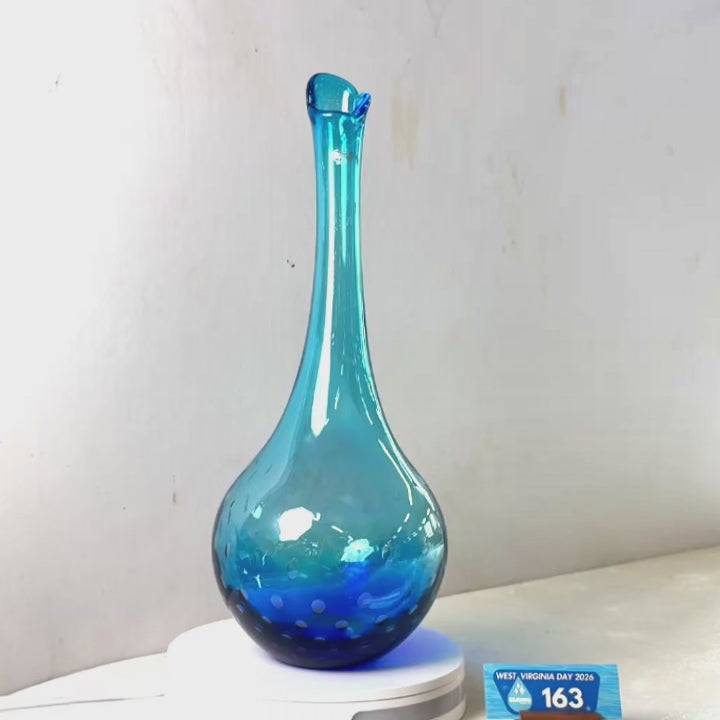 A translucent blue glass vase with a flared rim and dotted pattern near the base, displayed on a white circular stand against a plain light gray wall.