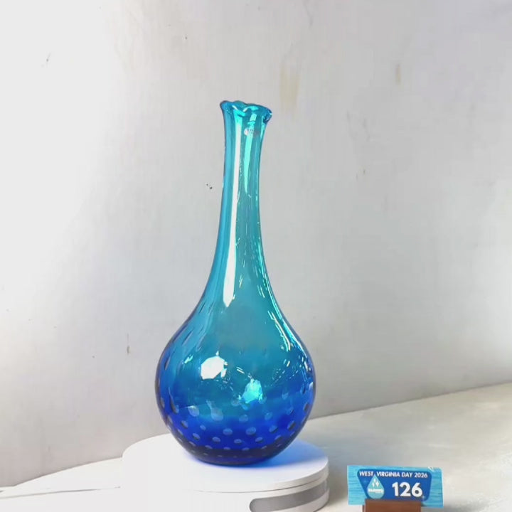 A translucent blue glass vase with a flared neck and dotted pattern near the base, displayed on a white circular light box. A small wrapped item and a brown block are placed beside it on a white surface against a plain light gray wall.