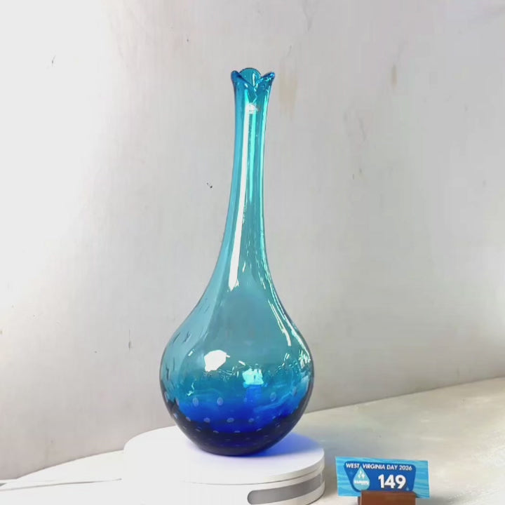 A translucent blue glass vase with a flared rim and dotted pattern near the base, displayed on a white circular stand against a plain light gray wall.