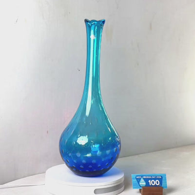 A translucent blue glass vase with a flared rim and dotted pattern near the base, displayed on a white circular stand against a plain light gray wall.