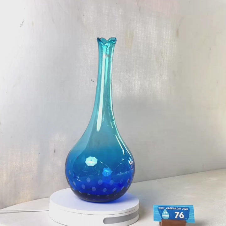 A translucent blue glass vase with a flared rim and subtle white speckled pattern near the base, displayed on a white round scale with a small price tag in front, against a plain light gray wall.
