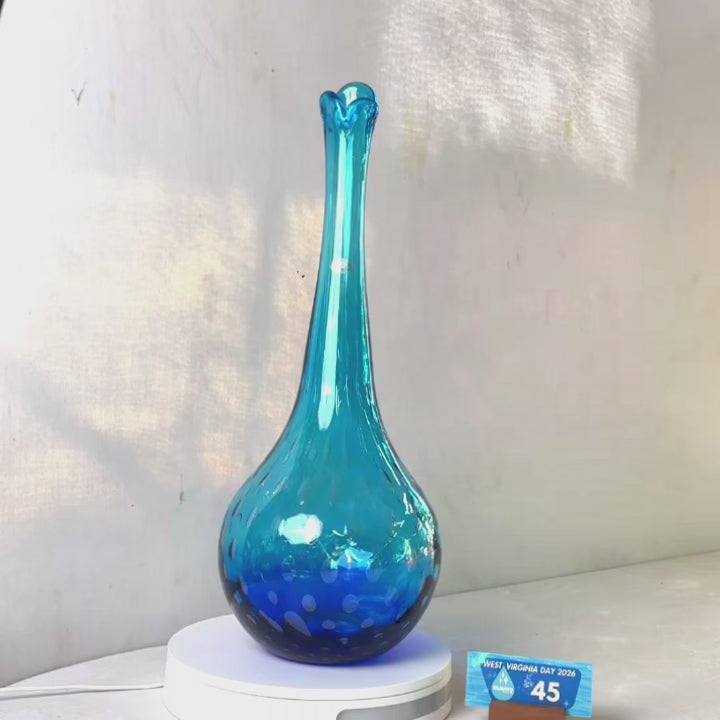 A translucent blue glass vase with a flared neck and dotted pattern, displayed on a white circular base against a light gray wall.