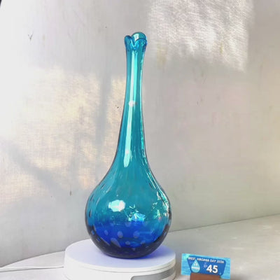 A translucent blue glass vase with a flared neck and dotted pattern, displayed on a white circular base against a light gray wall.