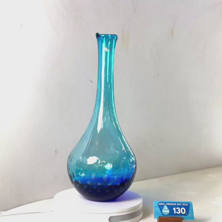 A translucent blue glass vase with a flared rim and dotted pattern near the base, displayed on a white circular stand against a plain light gray wall.