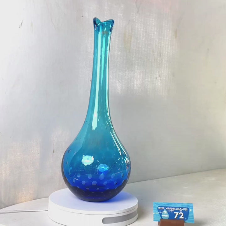 A translucent blue glass vase with a flared neck and dotted pattern, displayed on a white circular base against a light gray wall.