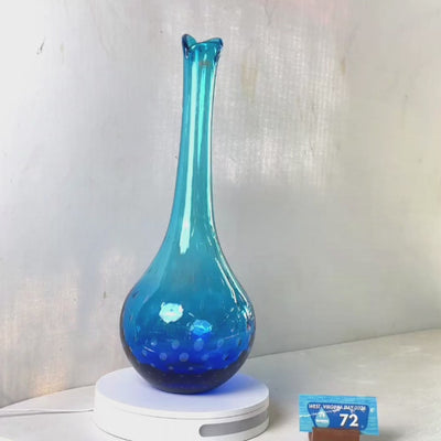 A translucent blue glass vase with a flared neck and dotted pattern, displayed on a white circular base against a light gray wall.