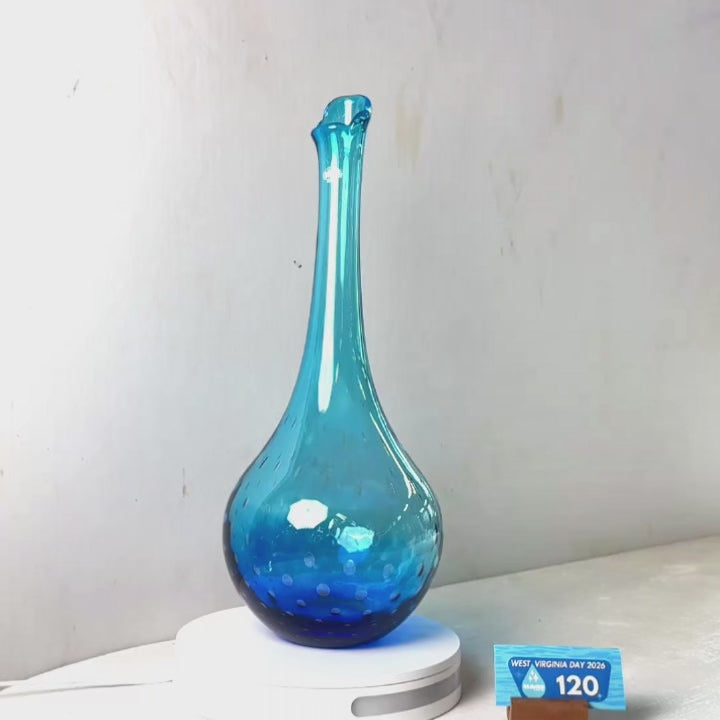 A translucent blue glass vase with a flared neck and dotted pattern near the base, displayed on a white circular light box. A small wrapped item and a brown block are placed beside it on a white surface against a plain light gray wall.