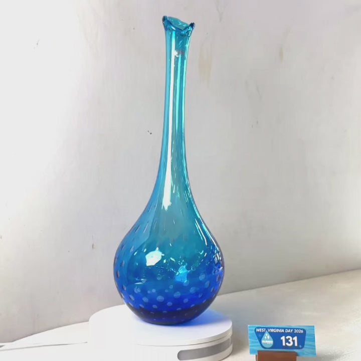A translucent blue glass vase with a flared rim and dotted pattern near the base, displayed on a white circular stand against a plain light gray wall.
