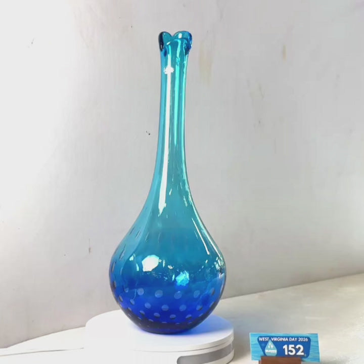 A translucent blue glass vase with a flared rim and dotted pattern near the base, displayed on a white circular stand against a plain light gray wall.