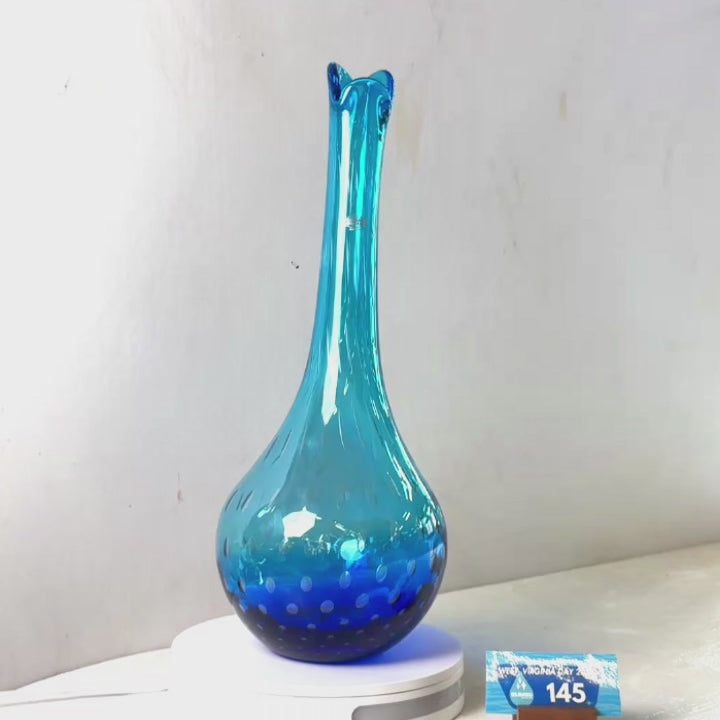 A translucent blue glass vase with a flared rim and dotted pattern near the base, displayed on a white circular stand against a plain light gray wall.