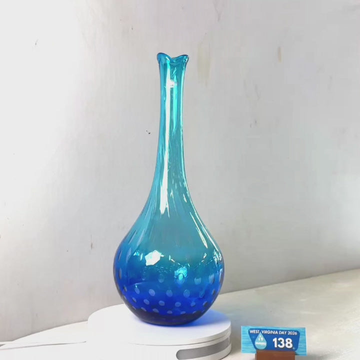 A translucent blue glass vase with a flared rim and dotted pattern near the base, displayed on a white circular stand against a plain light gray wall.