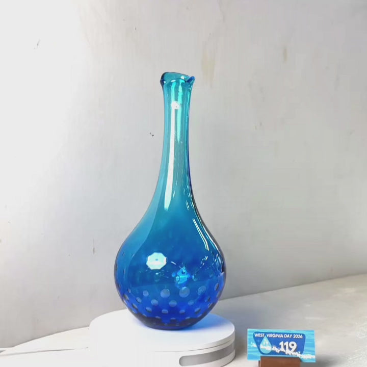 A translucent blue glass vase with a flared neck and dotted pattern near the base, displayed on a white circular light box. A small wrapped item and a brown block are placed beside it on a white surface against a plain light gray wall.