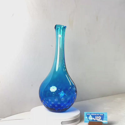 A translucent blue glass vase with a flared neck and dotted pattern near the base, displayed on a white circular light box. A small wrapped item and a brown block are placed beside it on a white surface against a plain light gray wall.