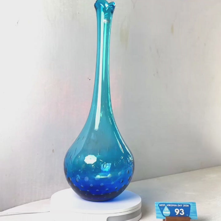 A translucent blue glass vase with a flared rim and subtle white speckled pattern near the base, displayed on a white round scale with a small price tag in front, against a plain light gray wall.