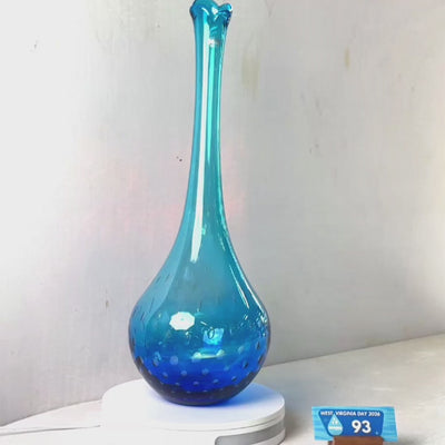 A translucent blue glass vase with a flared rim and subtle white speckled pattern near the base, displayed on a white round scale with a small price tag in front, against a plain light gray wall.