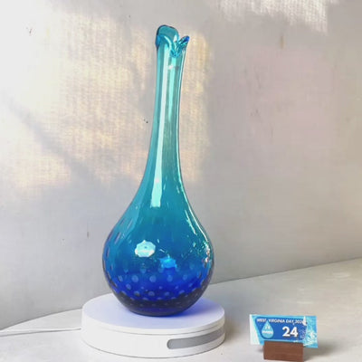 A translucent blue glass vase with a flared neck and dotted pattern, displayed on a white circular base against a light gray wall.