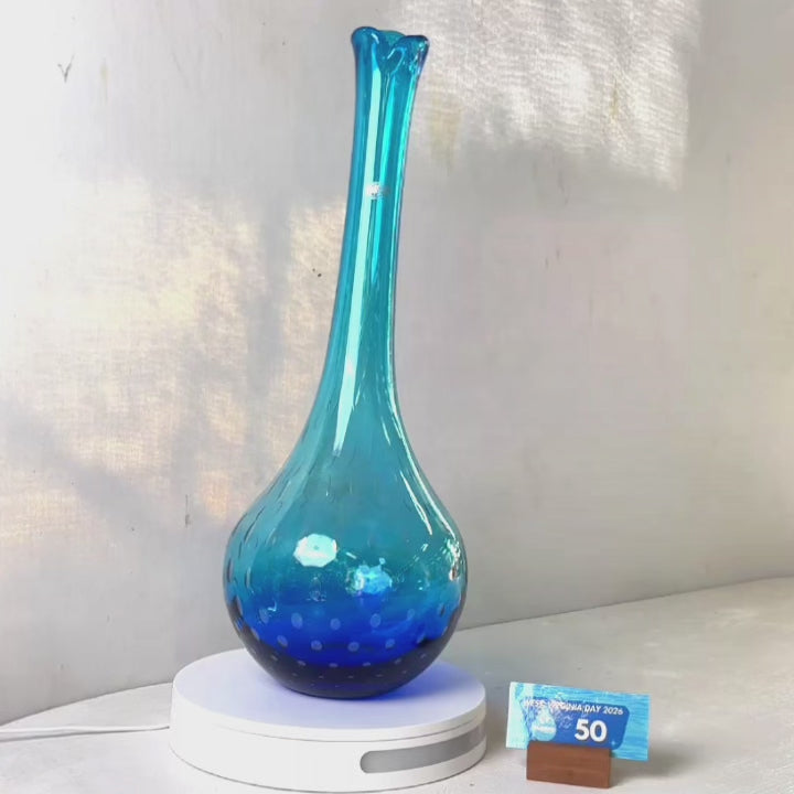 A translucent blue glass vase with a flared neck and dotted pattern, displayed on a white circular base against a light gray wall.