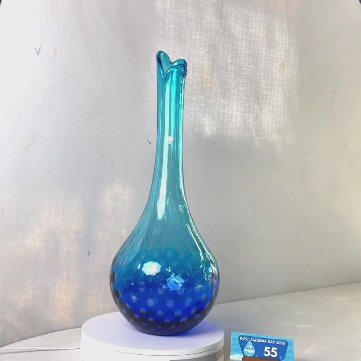 A translucent blue glass vase with a flared neck and dotted pattern, displayed on a white circular base against a light gray wall.
