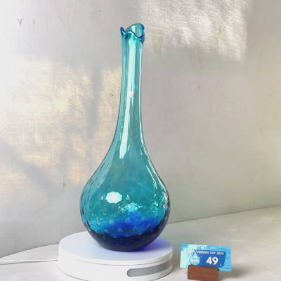 A translucent blue glass vase with a flared neck and dotted pattern, displayed on a white circular base against a light gray wall.