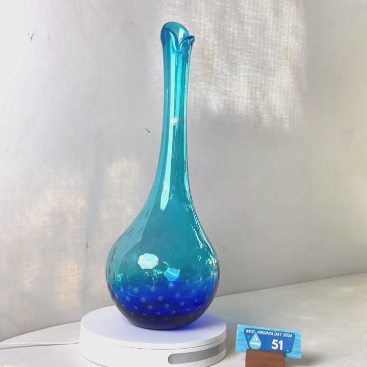 A translucent blue glass vase with a flared neck and dotted pattern, displayed on a white circular base against a light gray wall.