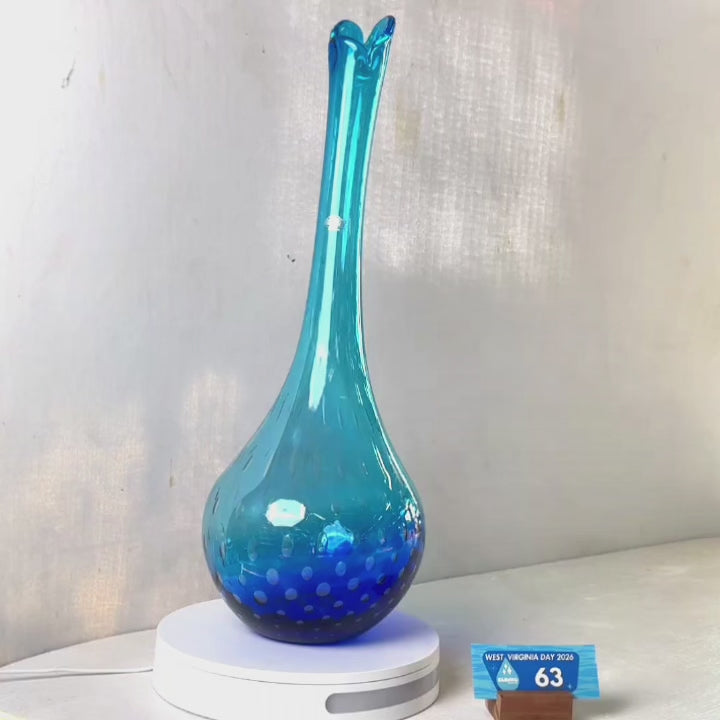 A translucent blue glass vase with a flared neck and dotted pattern, displayed on a white circular base against a light gray wall.